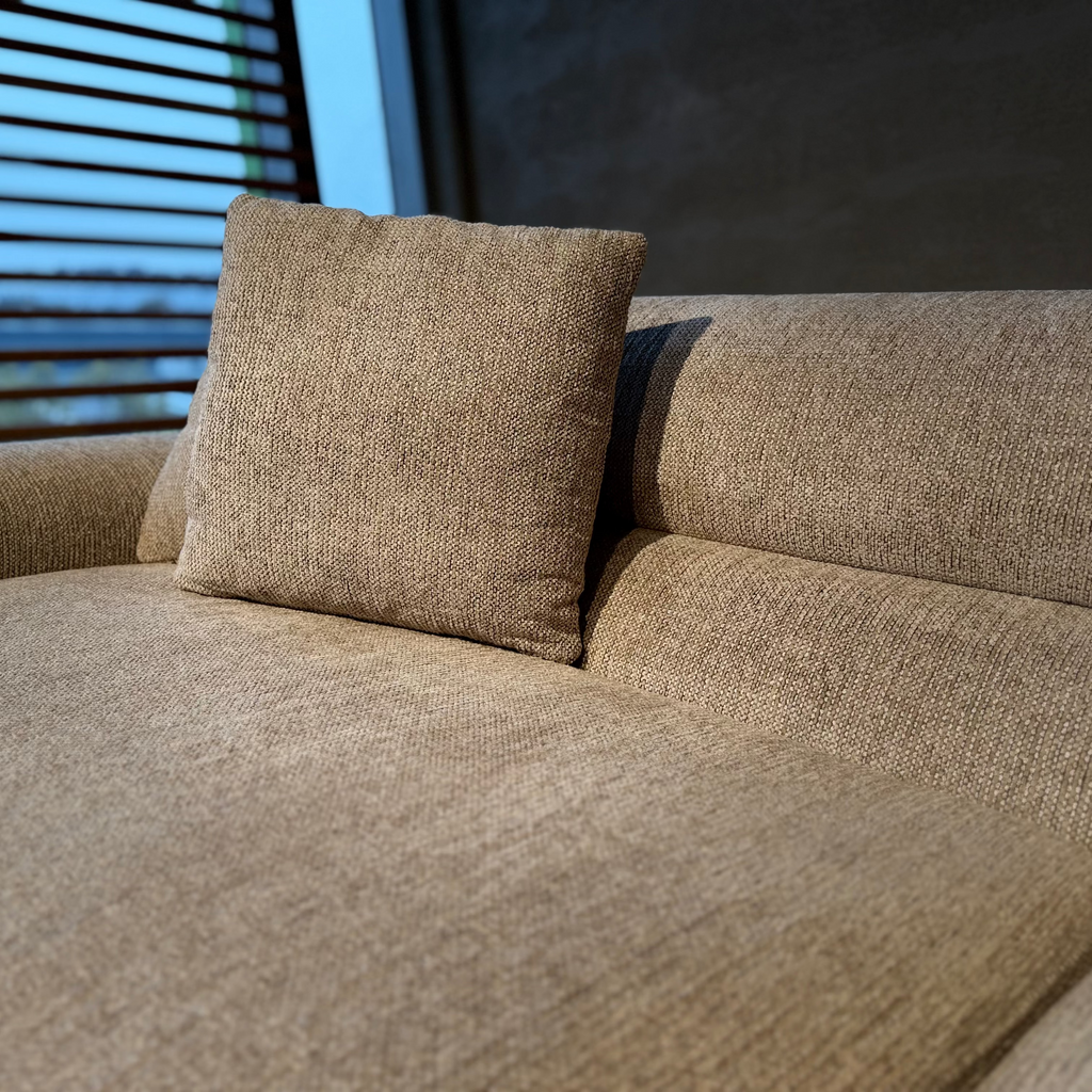 A close-up of a beige textured fabric sofa with a matching square throw pillow, set against a dark wall and window blinds.