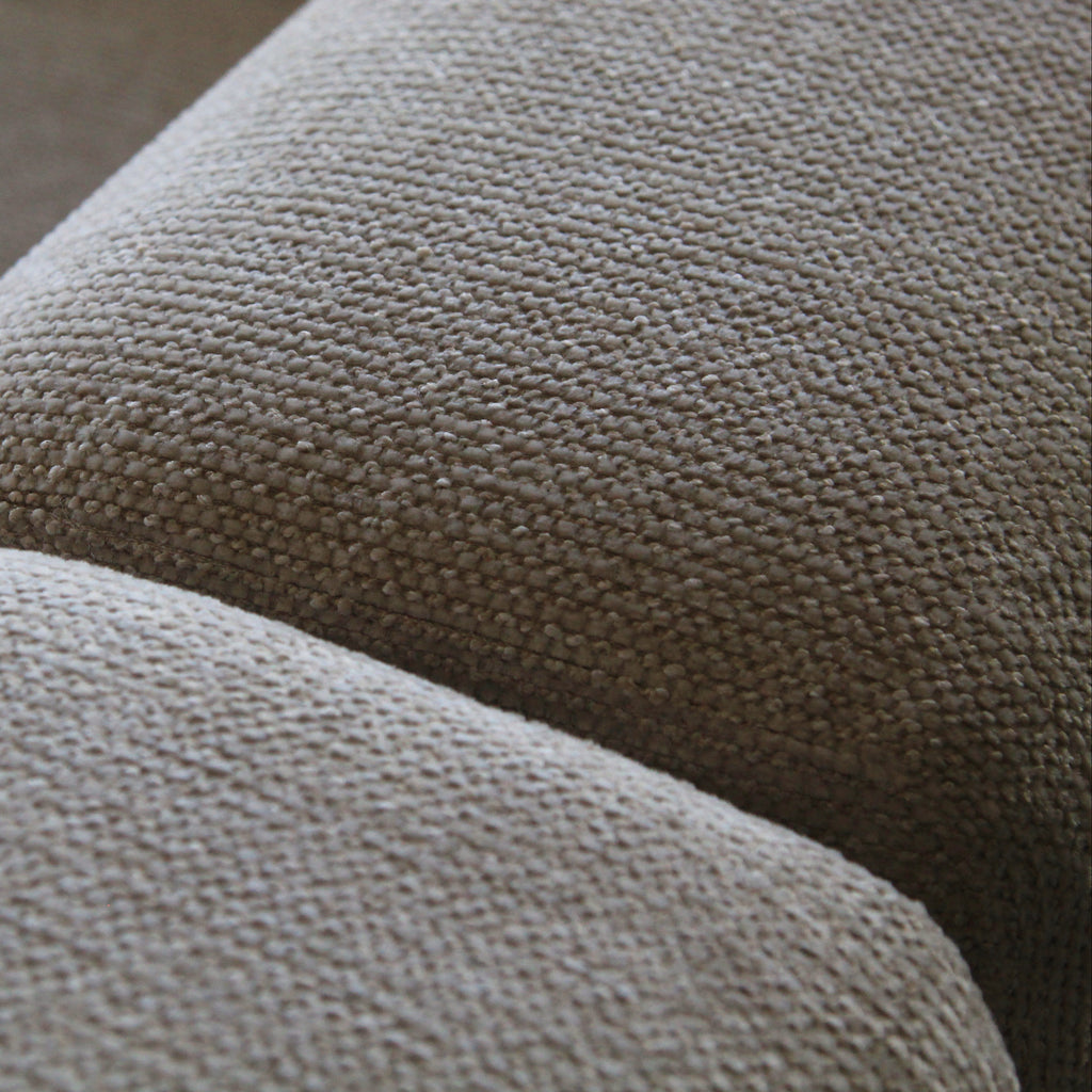 Close-up of a textured woven fabric upholstery in a warm taupe-beige color, showing the detailed weave and curved contours of what appears to be a sofa or cushion.