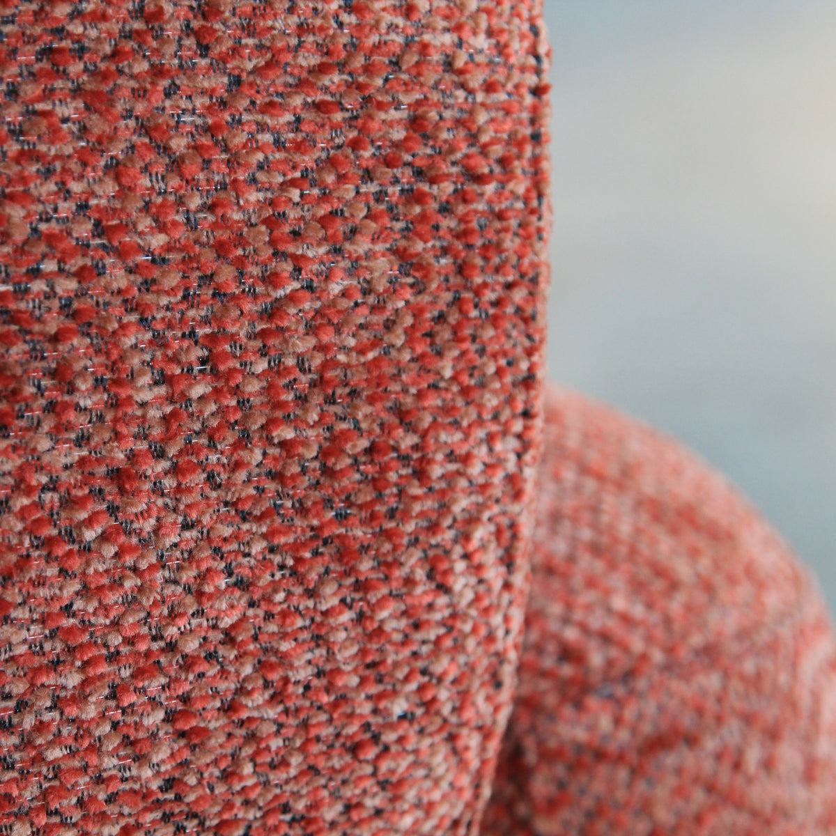 Close-up of a textured woven fabric in a heathered rust red, orange, and black pattern, with a blurred light gray background.