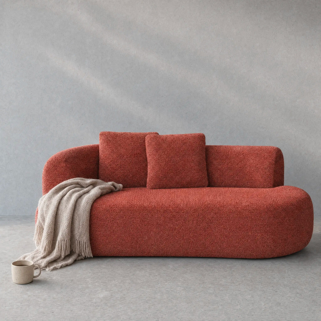 A modern curved sofa upholstered in a textured terracotta fabric, styled with two matching back cushions and a draped beige throw blanket, set against a textured gray background.