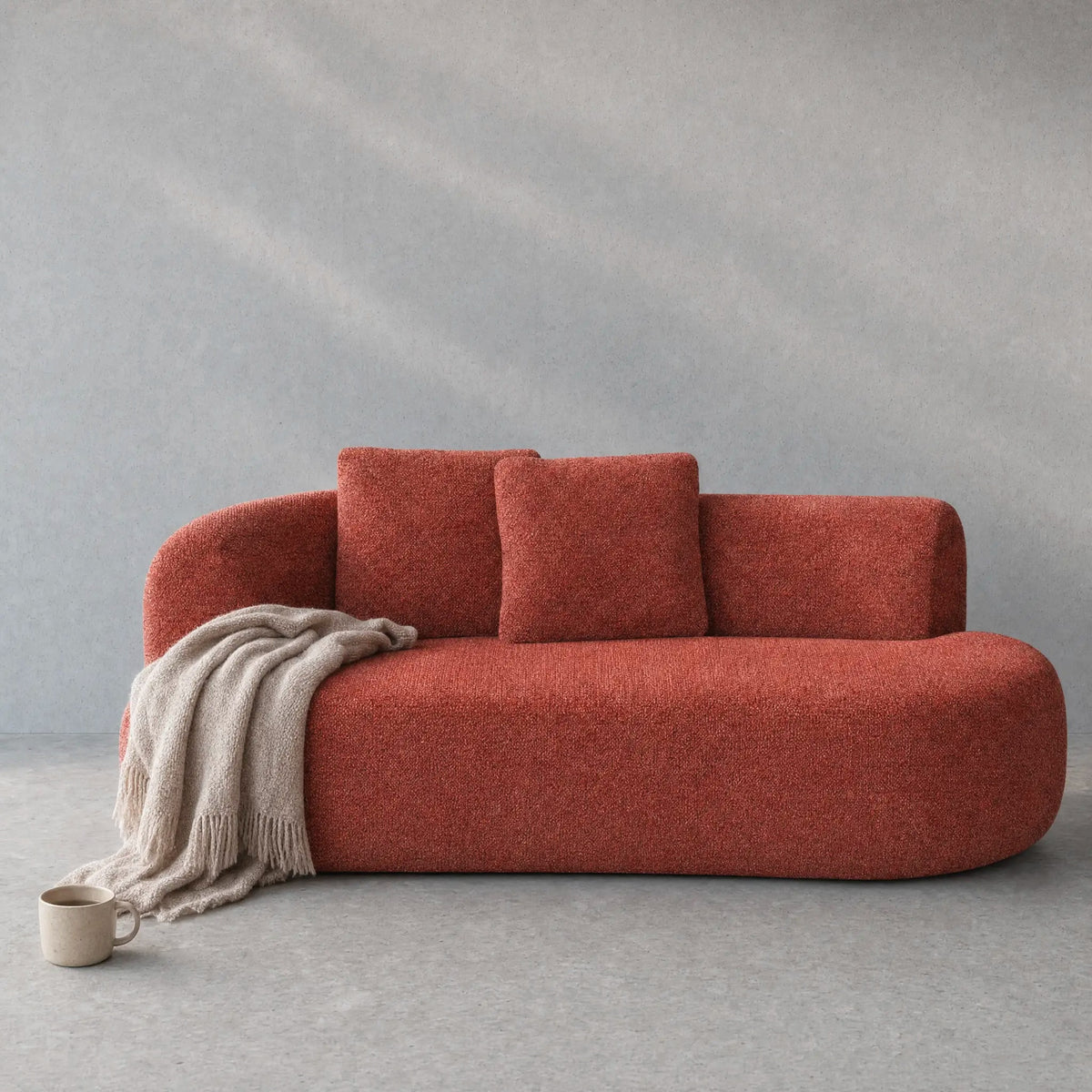 A modern curved sofa upholstered in a textured terracotta fabric, styled with two matching back cushions and a draped beige throw blanket, set against a textured gray background.
