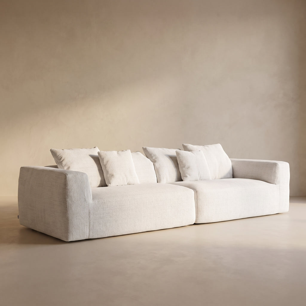 A modern low-profile sofa upholstered in a light cream fabric with matching throw pillows, set against a minimalist beige plaster wall and floor.