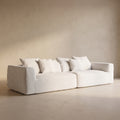 A modern low-profile sofa upholstered in a light cream fabric with matching throw pillows, set against a minimalist beige plaster wall and floor.